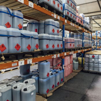 A shot of shelving units filled with plastic containers of ink in an ink factory. The space is an industrial warehouse, and there are more containers of ink wrapped in plastic on pallets beside the shelving units.