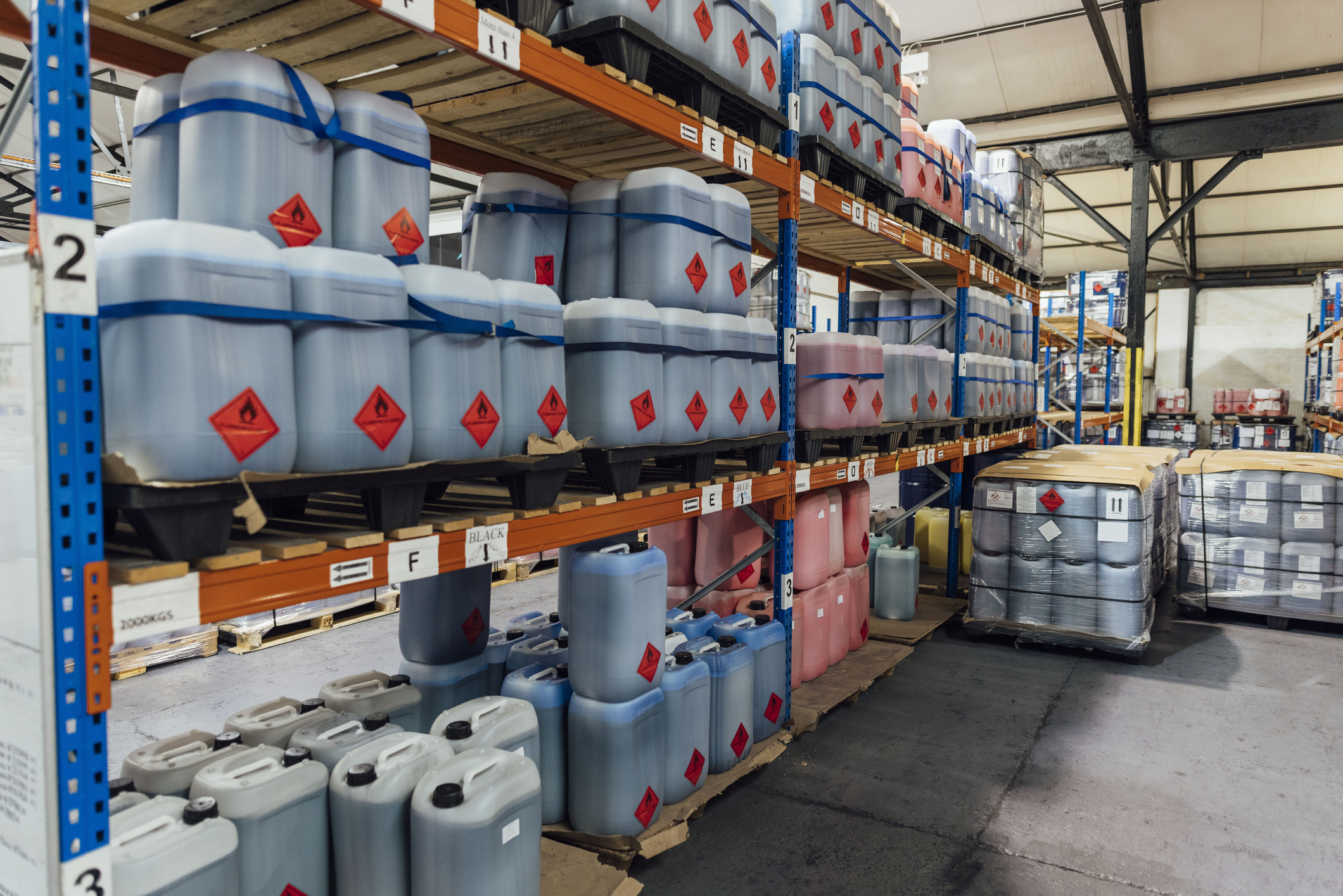 A shot of shelving units filled with plastic containers of ink in an ink factory. The space is an industrial warehouse, and there are more containers of ink wrapped in plastic on pallets beside the shelving units.